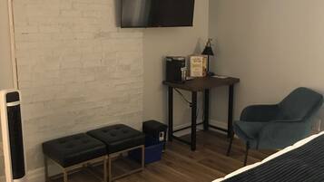 A room with a bed, a bench, a table with a coffee maker, a chair, and a TV mounted on the wall.