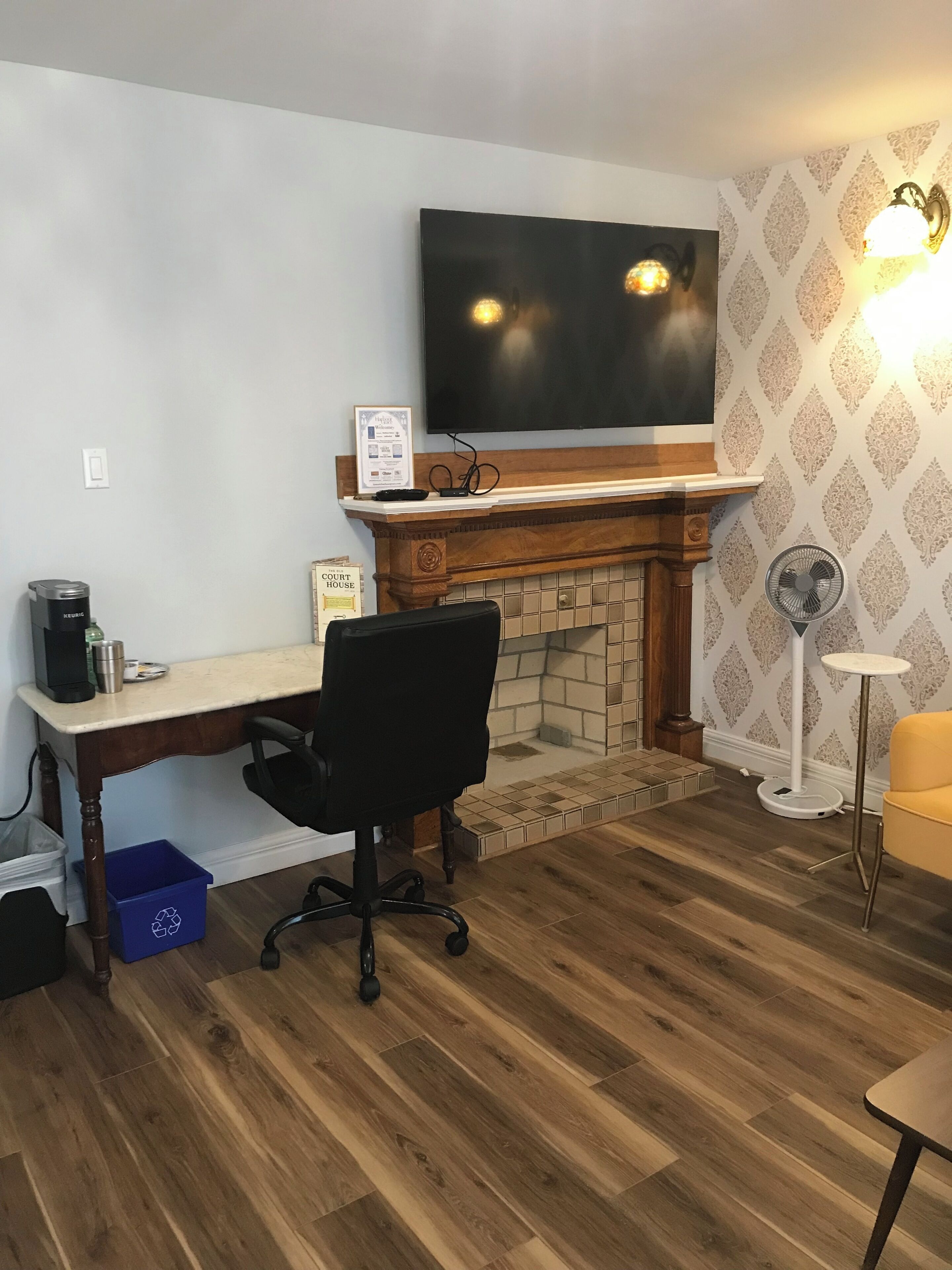 A room with a fireplace, a television mounted above it, a desk with a coffee maker, a black office chair, and a floor fan.