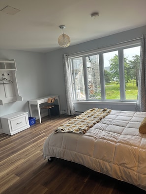 A bedroom with a large bed, a desk, and a chair. There is a window with a view of greenery outside.
