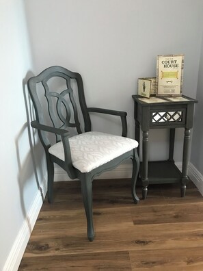 Una silla de madera oscura con asiento y respaldo acolchados, ubicada al lado de una pequeña mesa auxiliar de madera con una placa y un cuadro enmarcado.