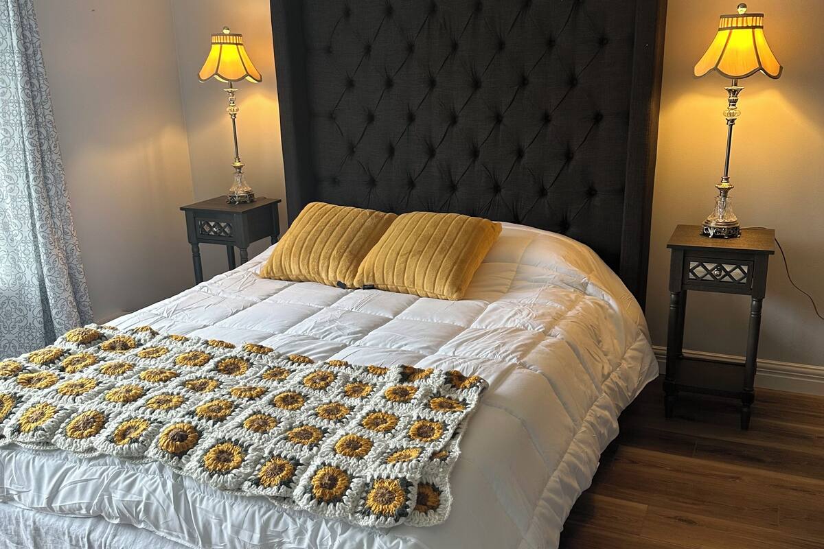 Una cama cuidadosamente tendida con un edredón blanco y una colcha de ganchillo con motivos de girasoles. Hay dos mesitas de noche con lámparas, un cabecero tapizado oscuro y suelo de madera.