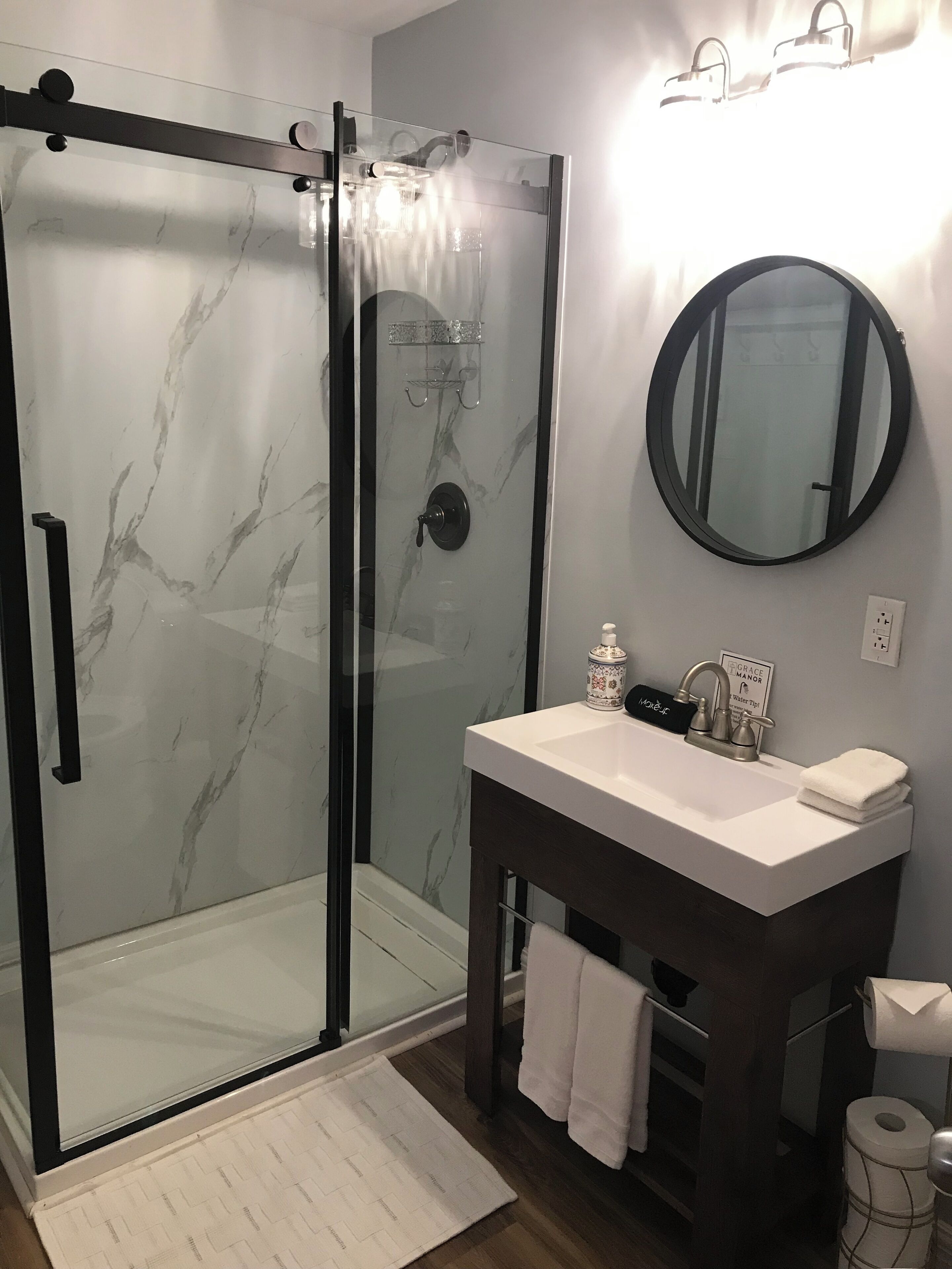Une salle de bain moderne avec une douche à parois de verre, un lavabo blanc et un meuble-vasque foncé, ainsi qu’un miroir rond.