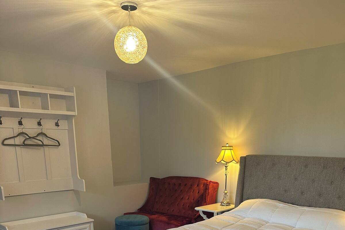 Un dormitorio con una cama grande, una lámpara colgante, una mesita de noche con lámpara, una silla roja y un mueble blanco para guardar objetos.