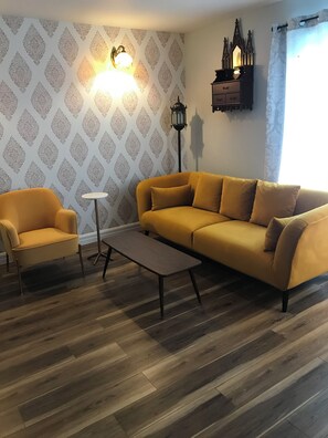 Ett vardagsrum med en mönstrad tapet, en soffa, en fåtölj, ett litet bord och en golvlampa.