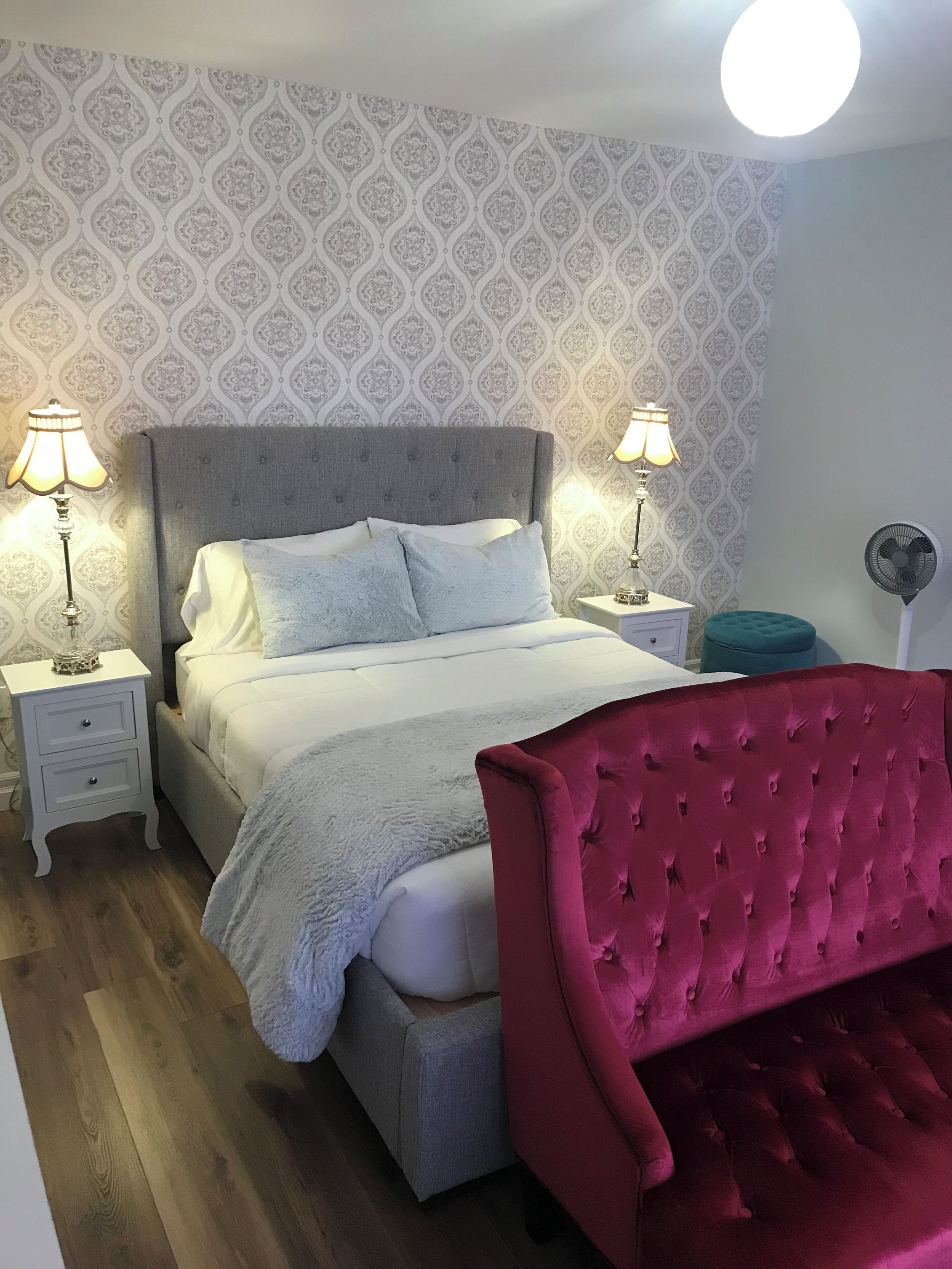 A bedroom with a bed, two bedside lamps, a nightstand, a red upholstered chair, and a wall with a patterned wallpaper.