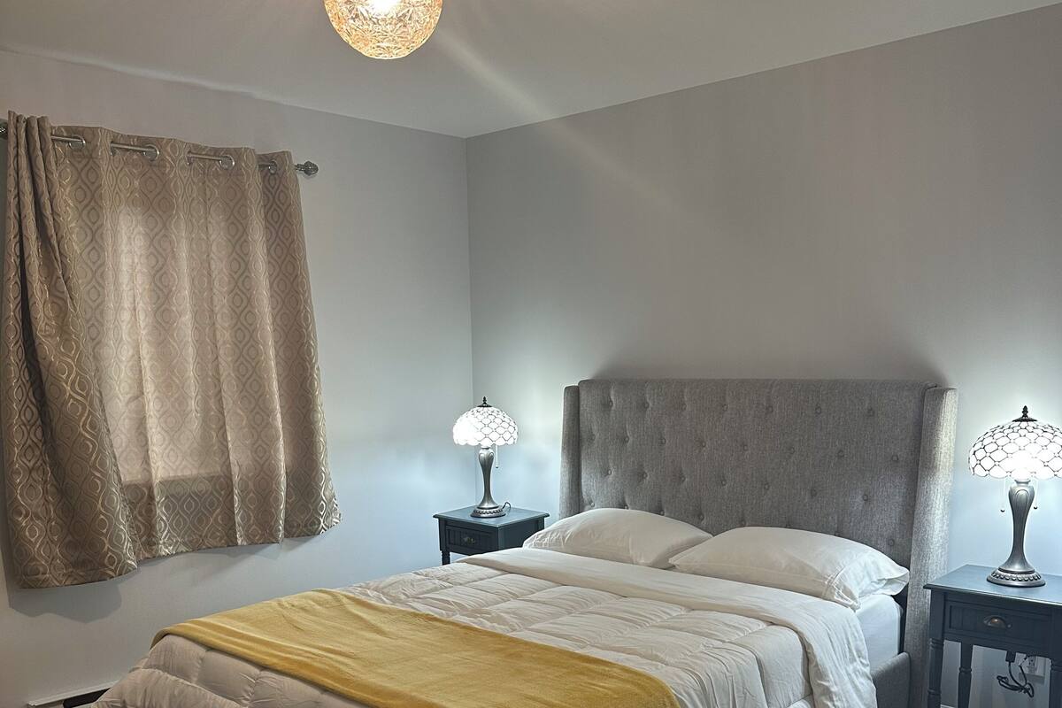 Un dormitorio con una cama, dos mesitas de noche, un banco, una lámpara colgante y una ventana con cortinas.