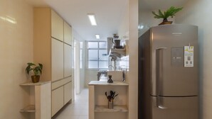A modern kitchen with a stainless steel refrigerator, a built-in oven, and a sink. There is a potted plant on the countertop and a shelf with kitchen utensils.