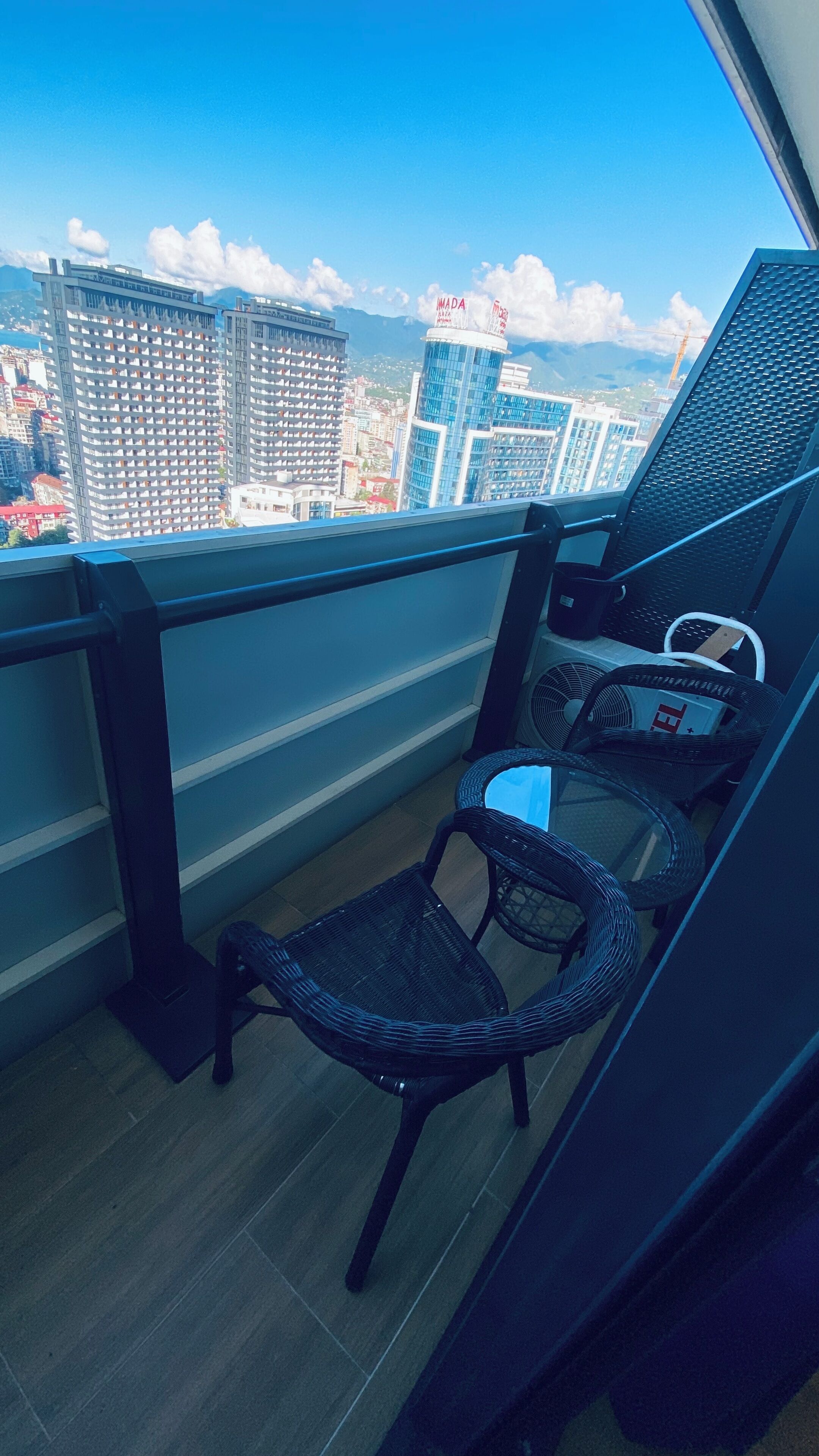 A balcony with a wicker chair, a small table, and a fan. The view includes high-rise buildings and a clear sky.