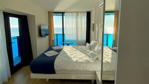 A modern hotel room with a large bed, a TV, and a balcony with a view of the sea.