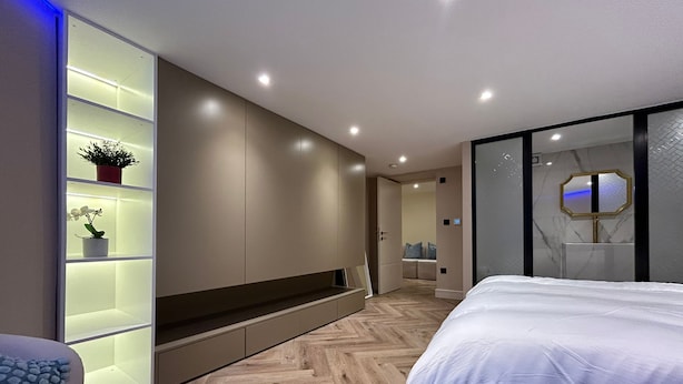 Ein modernes Schlafzimmer mit einem großen Bett, einem eingebauten Kleiderschrank mit beleuchteten Regalen und einem Holzboden mit Fischgrätmuster.