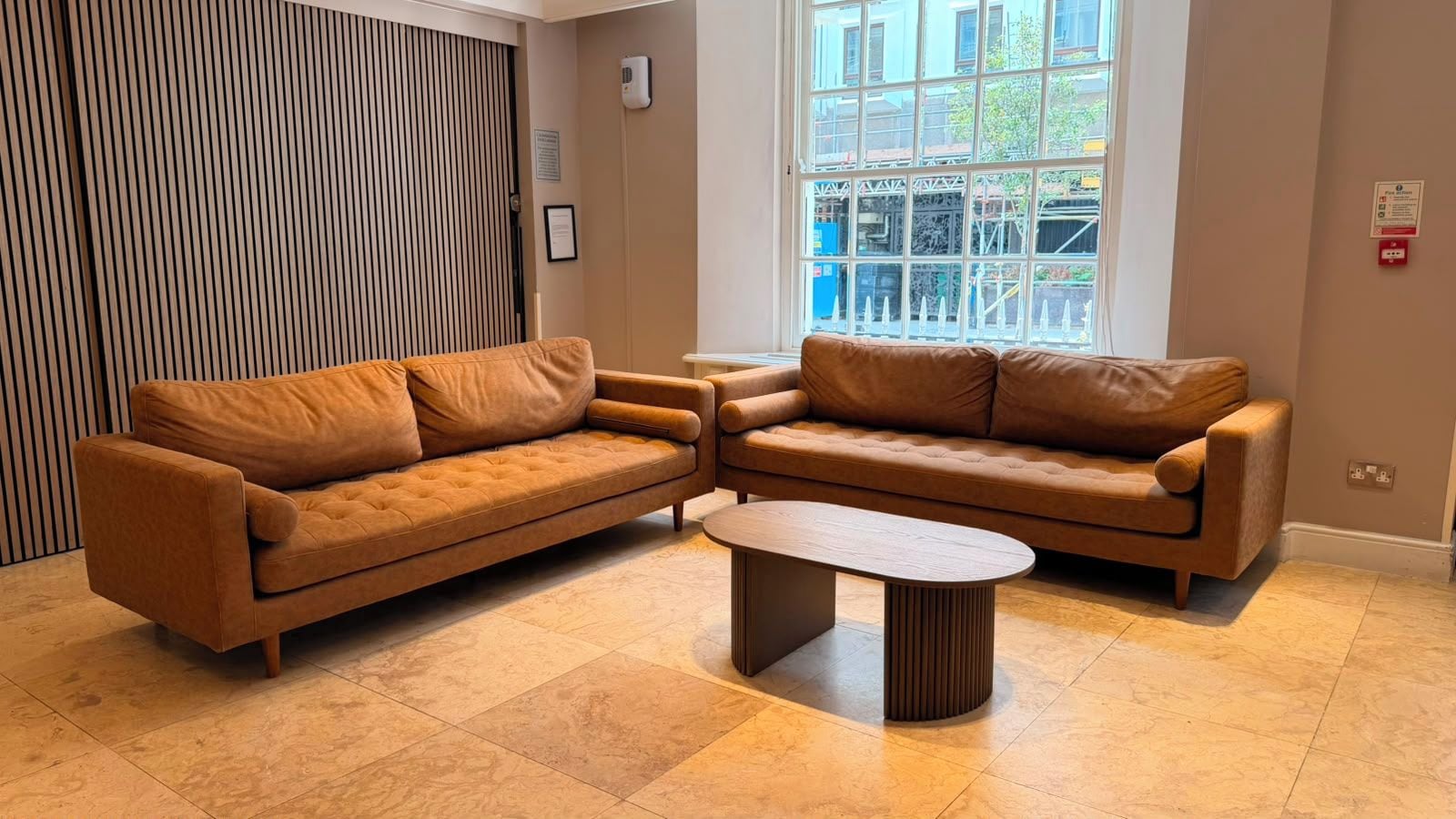 Un salon moderne avec deux canapés marron, une table basse en bois et une grande fenêtre donnant sur des immeubles.