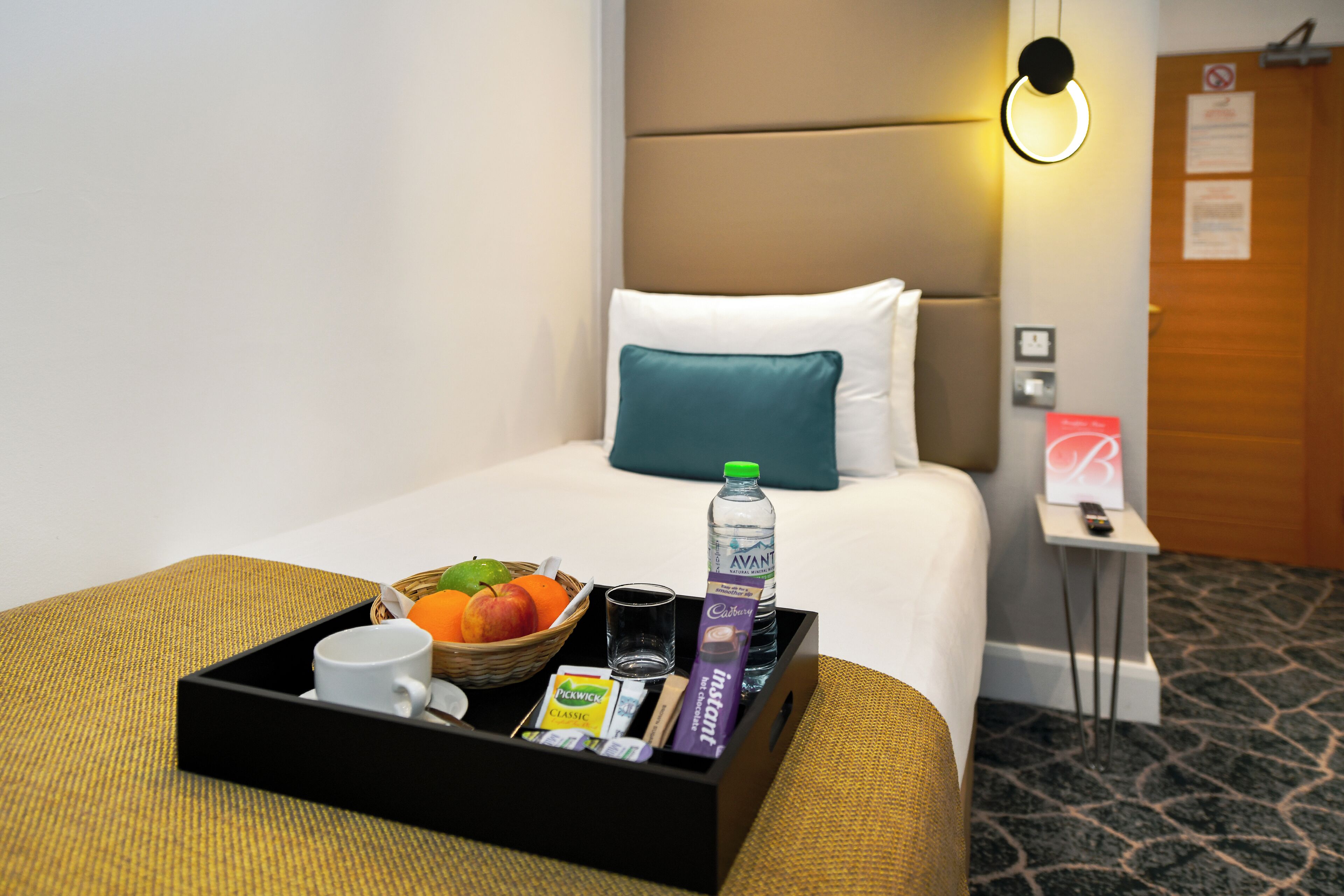 A hotel room with a neatly made bed, a bedside table with a remote control, and a tray with breakfast items including fruits, a bottle of water, and a cup.