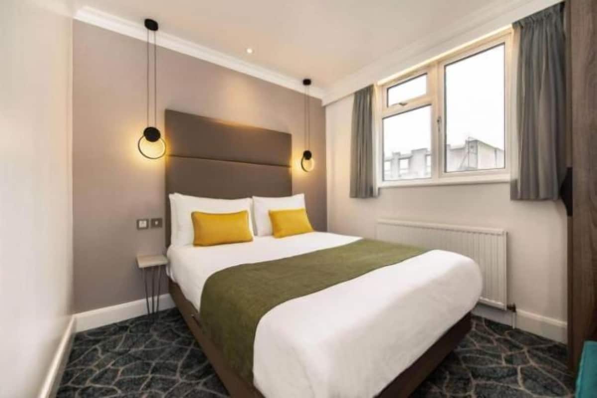 A hotel room with a large bed, a nightstand, a window with curtains, and a wall-mounted light fixture.