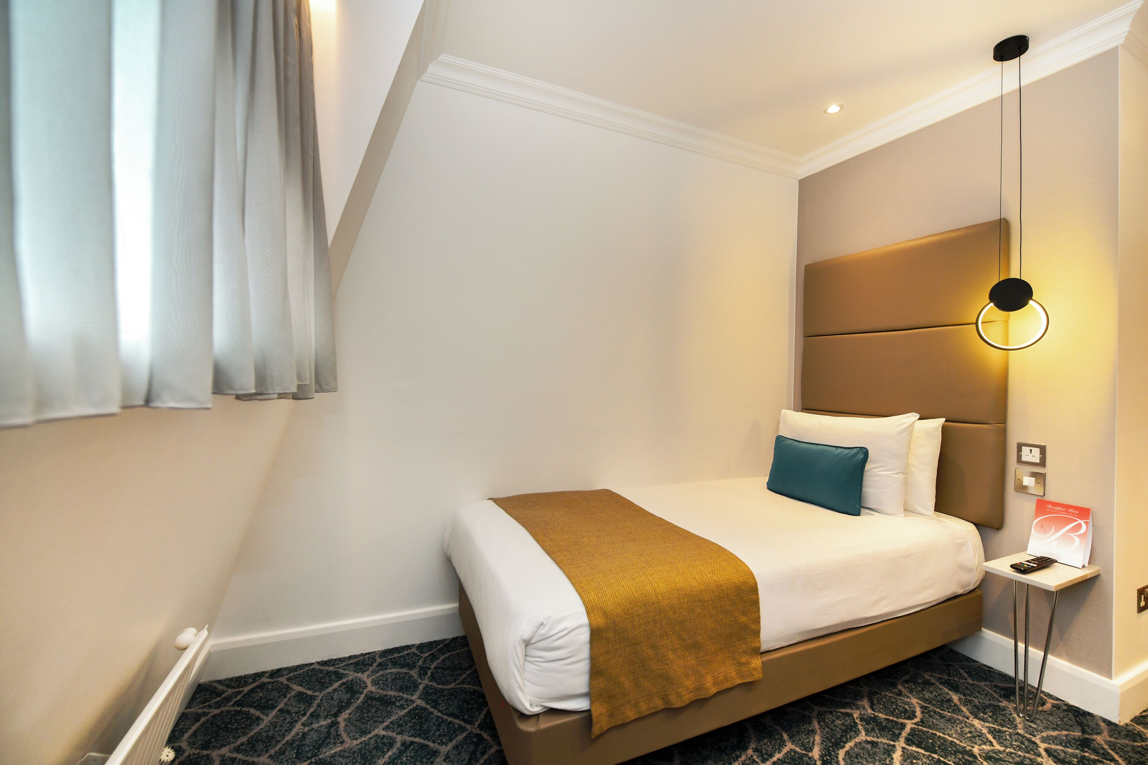 A hotel room with a bed, a bedside table, a nightstand, a lamp, and a window with curtains.