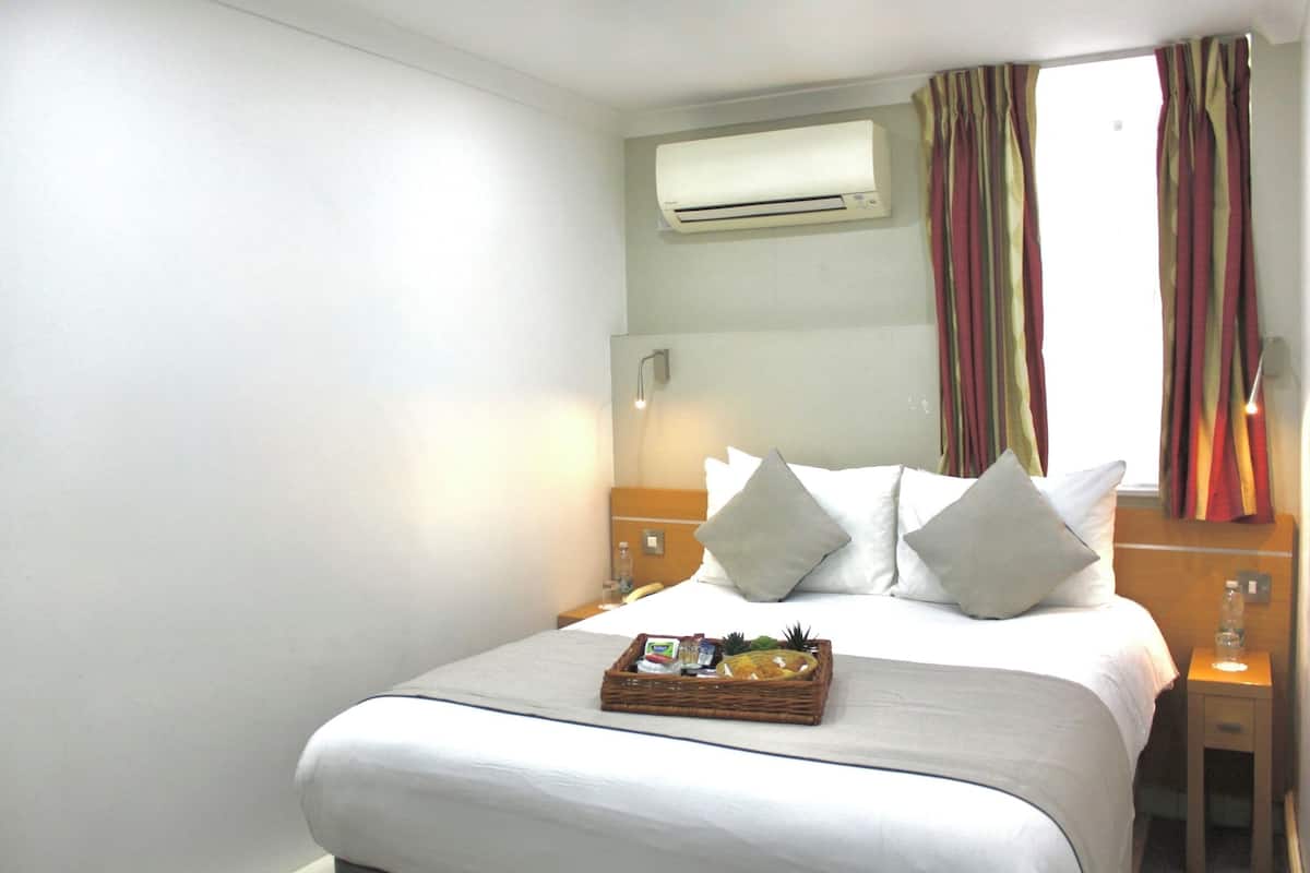 A hotel room with a neatly made bed, a bedside table with a lamp, a wall-mounted air conditioner, and a window with curtains.
