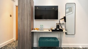 Een moderne hotelkamer met een grote flatscreen televisie aan de muur, een klein bureau met spiegel, een blauwgroene bankje en een kamerbreed tapijt met patroon.