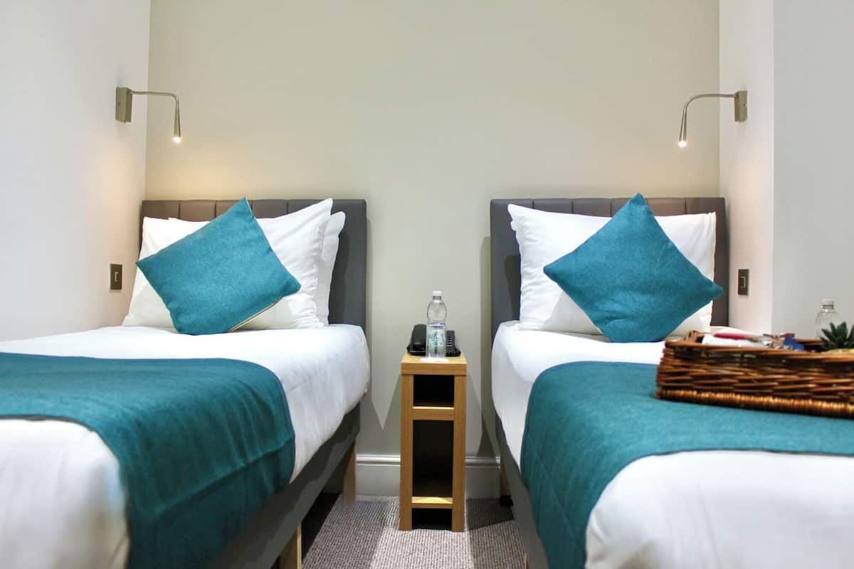A hotel room with two beds, each with a teal bedspread and pillows, a small wooden nightstand with a bottle and a phone, and a wicker basket on the right side.