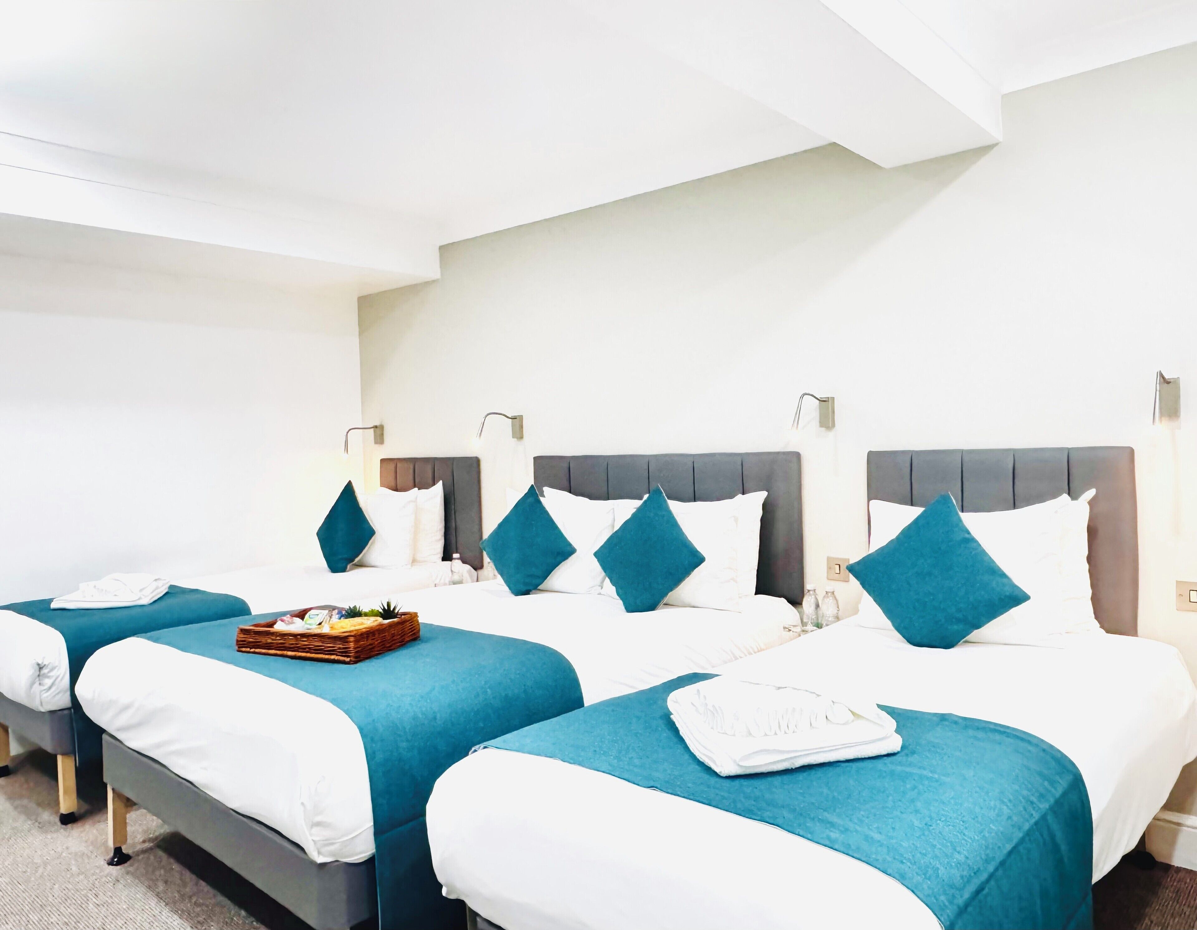Ein Hotelzimmer mit zwei Betten, jedes mit einer blauen Tagesdecke und weißer Bettwäsche. Zwei Nachttische mit Lampen stehen bereit, und auf einem Bett liegt ein kleiner Korb.