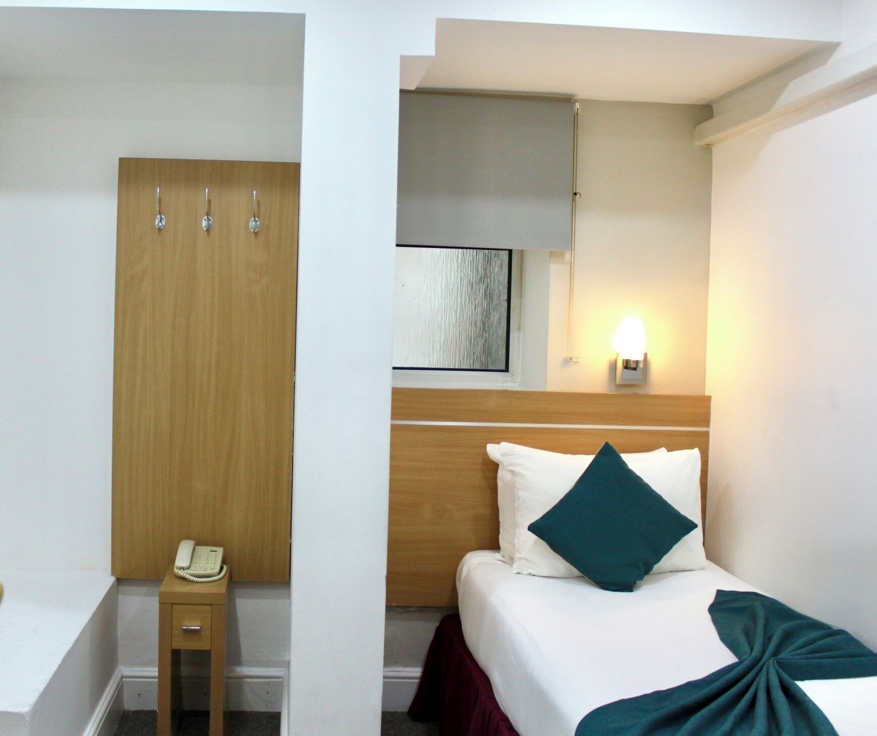 A hotel room with a bed, a bedside table with a telephone, a wooden wardrobe, and a wall-mounted light.
