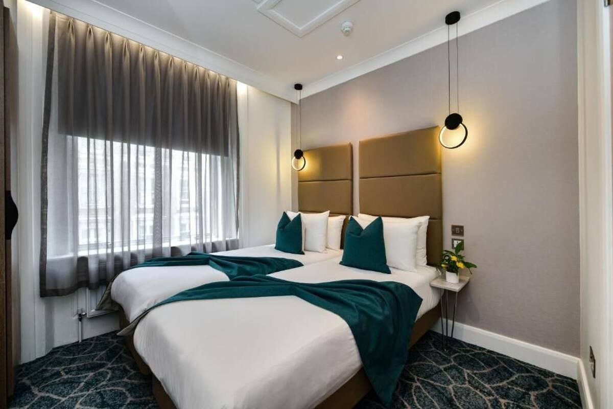 A hotel room with two beds, a large window with curtains, a headboard with integrated lighting, and a small bedside table with a plant.
