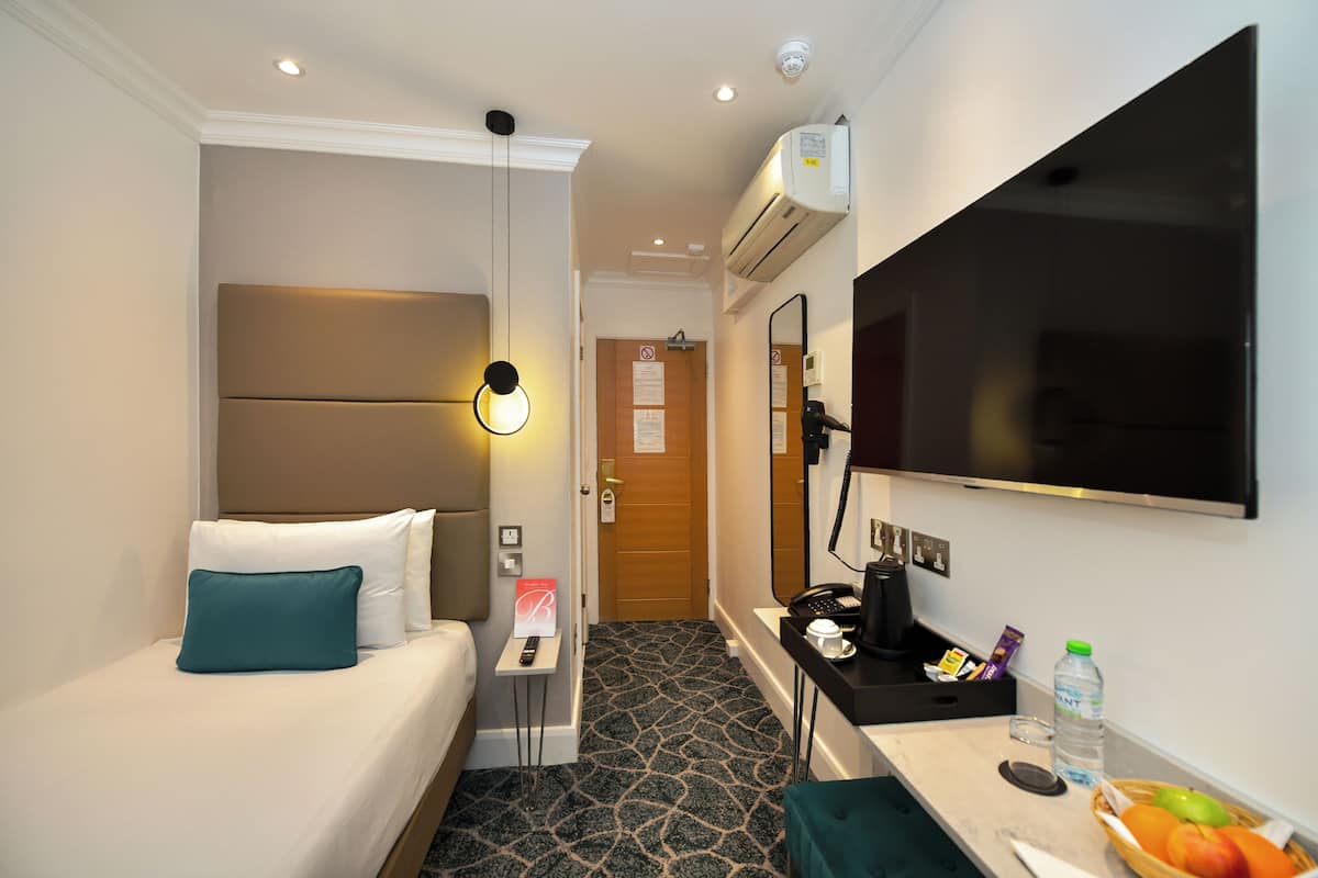 A hotel room with a bed, bedside table, a desk with a chair, a flat-screen TV, and a small kitchenette area.