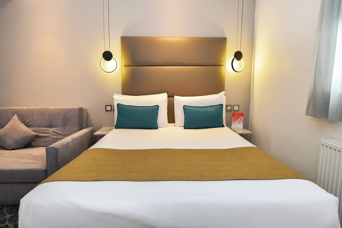 A hotel room with a large bed, two bedside tables, a nightstand, a sofa, and a wall-mounted TV.
