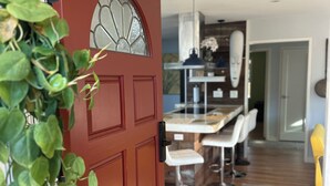 Una porta rossa con un vetro che dà su un'area pranzo con tavolo e sedie, un tappeto decorato e una ventola a soffitto.