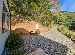 Un chemin de gravier mène à un banc en bois recouvert d’un tissu blanc, entouré de verdure et d’arbres.