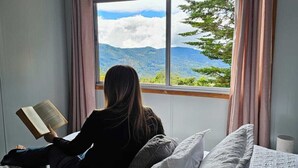 Iemand zit op een bed te lezen, met uitzicht op bergen en bomen.