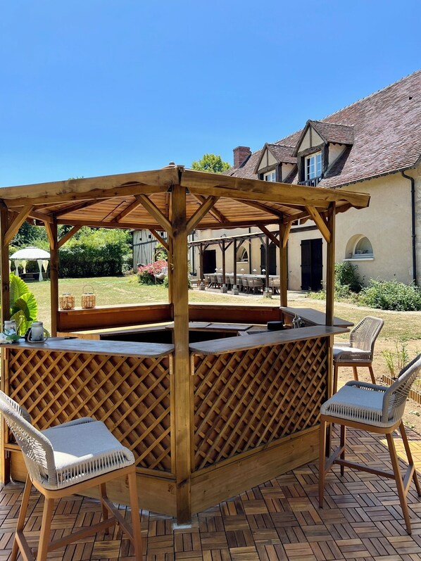 Una zona de bar exterior con barra de madera, un divisor de celosía, dos sillas acolchadas y un banco.
