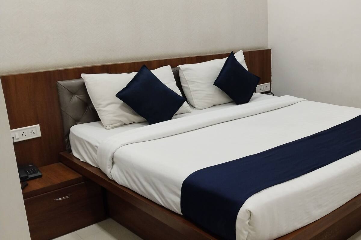Un lit bien fait, recouvert d’une couvre-lit blanche et bleu marine, avec une tête de lit en bois et une table de chevet surmontée d’une lampe.