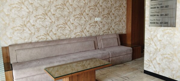 Une pièce avec un mur décoré d’un motif imitant le marbre, un canapé beige et une table avec un plateau en verre. Un panneau indique les numéros des chambres pour les différents étages.