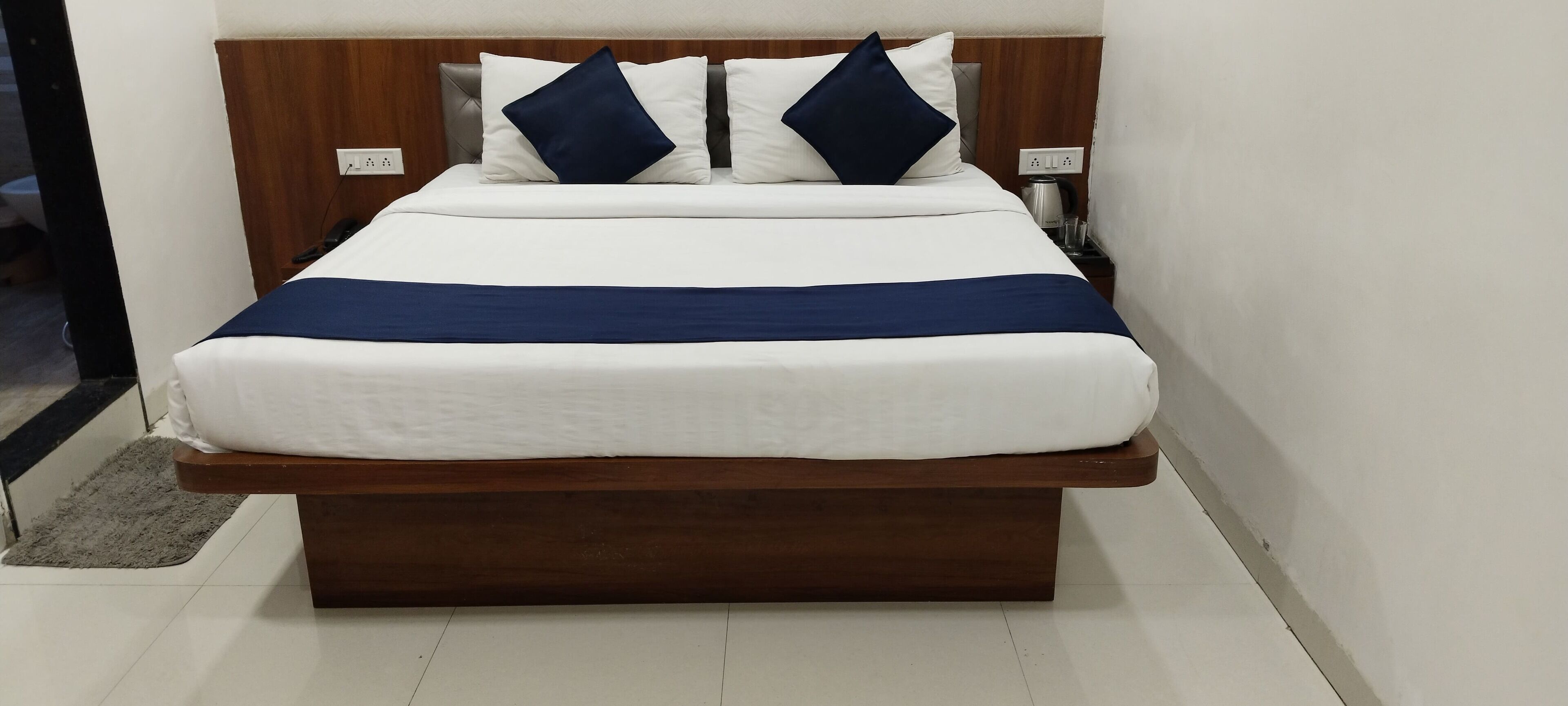 Un letto rifatto con cura, lenzuola bianche e due cuscini blu scuro, una testiera in legno e un comodino con una lampada e una presa di corrente.