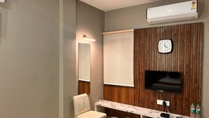 A hotel room with a wooden panel wall, a flat-screen TV mounted above a wooden desk, a chair, a small round table, and a wall-mounted clock. - Wishotel Inn Skymoon  (Indore)