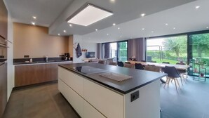 Eine moderne Küche mit einer zentralen Kochinsel, einem Essbereich mit Tisch und Stühlen sowie großen Fenstern mit Blick ins Freie.