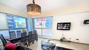 Una sala de estar moderna con un sofá de cuero, una mesa de comedor con sillas, una lámpara con estampado de cebra, un televisor de pantalla plana y ventanas grandes con vista a la ciudad.