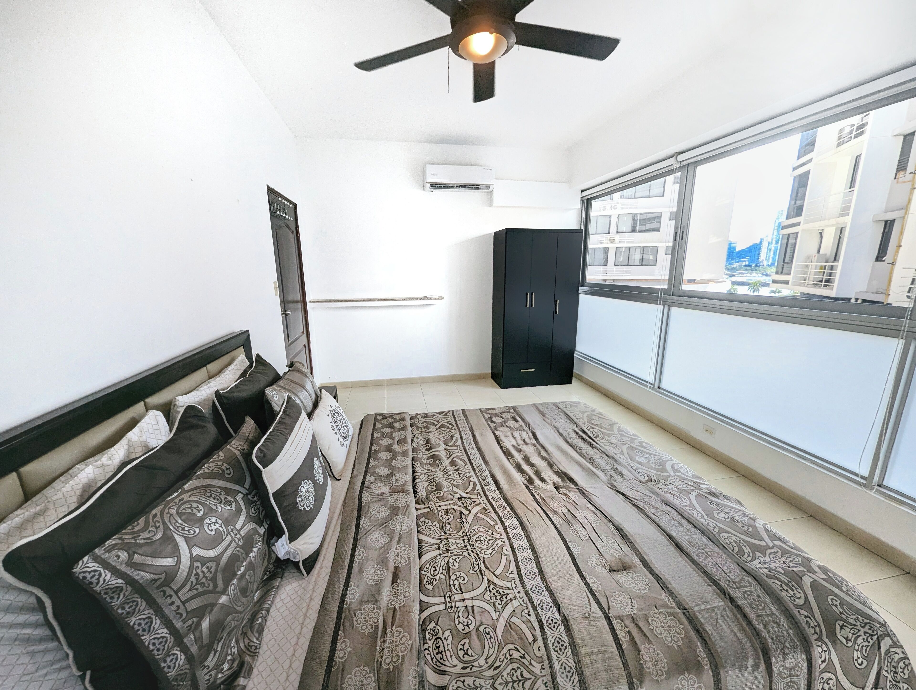 Una habitación con cama, ventilador de techo, aire acondicionado, armario y un amplio ventanal con vistas a edificios.