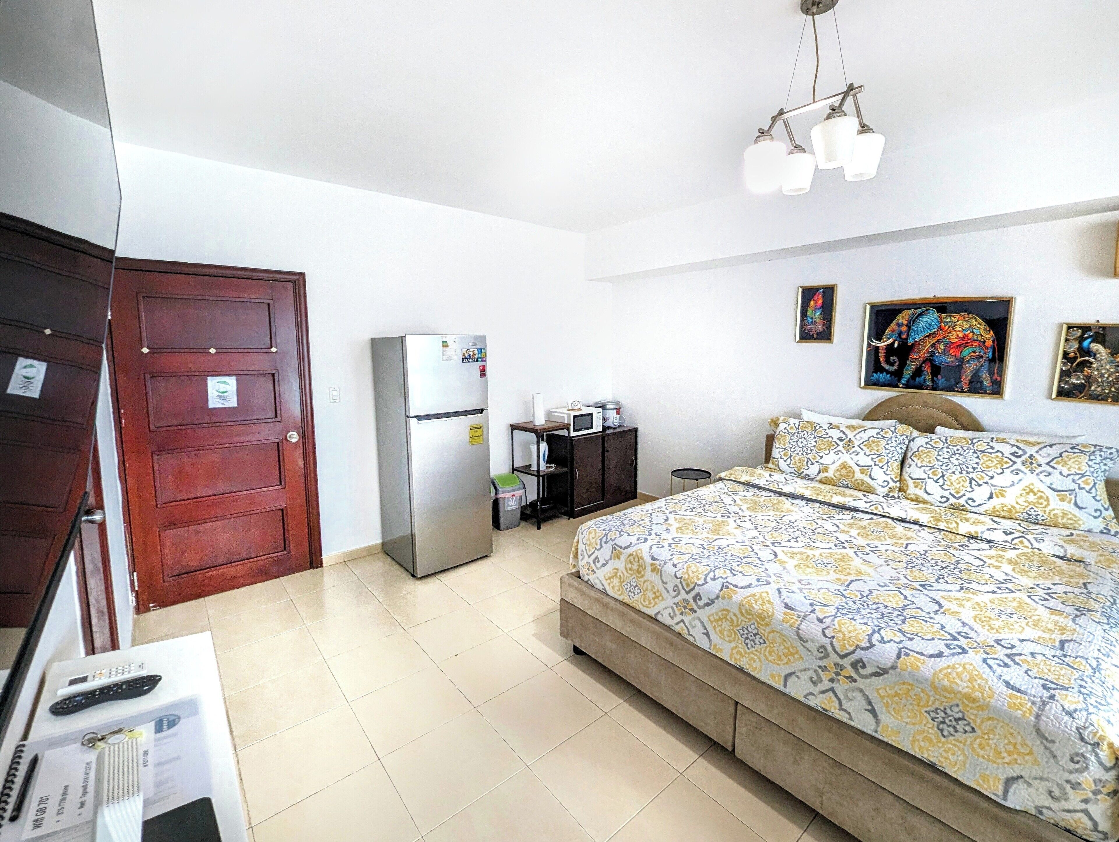 A bedroom with a bed, a refrigerator, a wooden door, a small table with a microwave, and a ceiling light fixture.