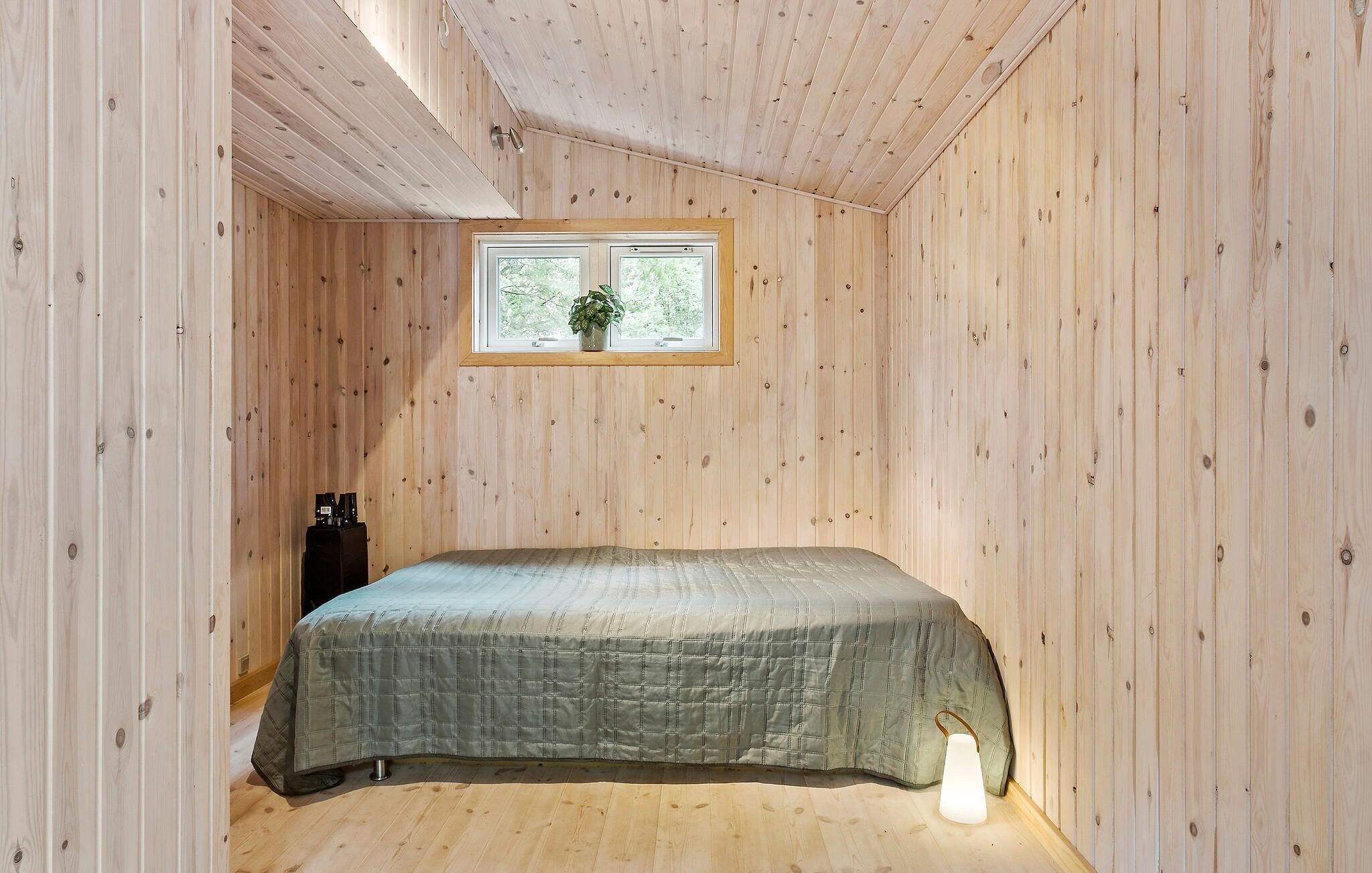 A small, cozy room with wooden walls and a single bed. There is a window with a view of greenery outside.