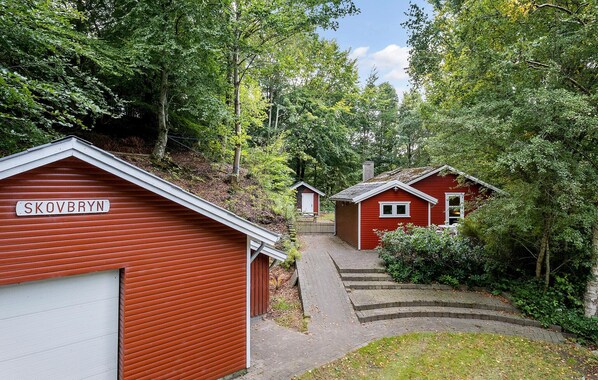 A red wooden cabin with a sign reading "SKOVBRYN" and a smaller adjacent cabin, surrounded by dense greenery. - Nice home in Gistrup with WiFi (Gistrup)