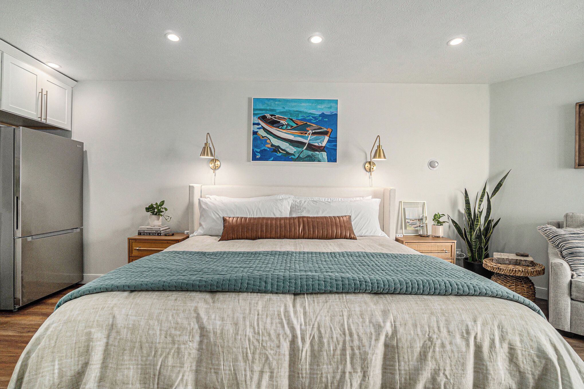 A bedroom with a bed, a painting of a boat, a refrigerator, bedside tables, and a plant.