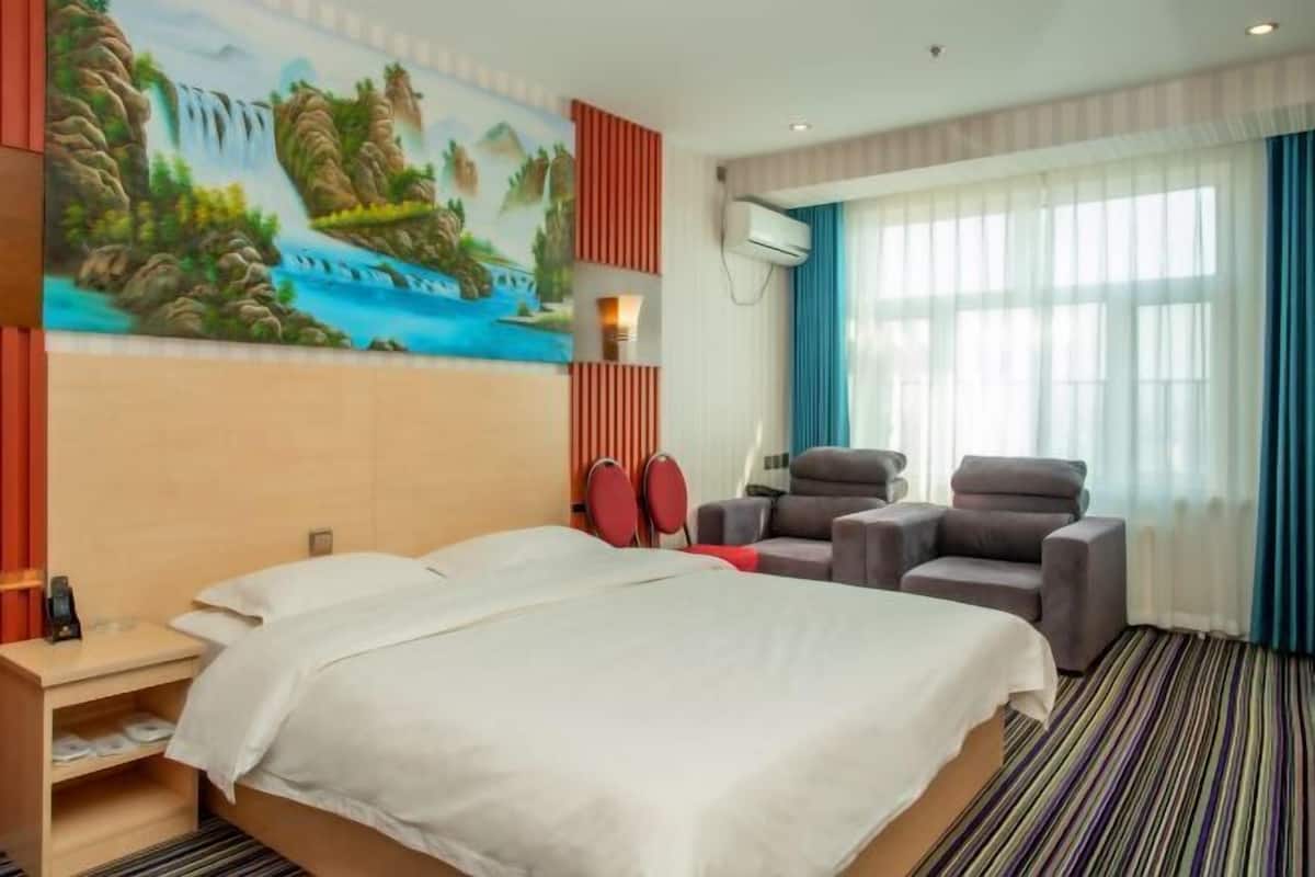 A hotel room with a bed, two armchairs, a painting, a window with curtains, and a wall-mounted air conditioner.
