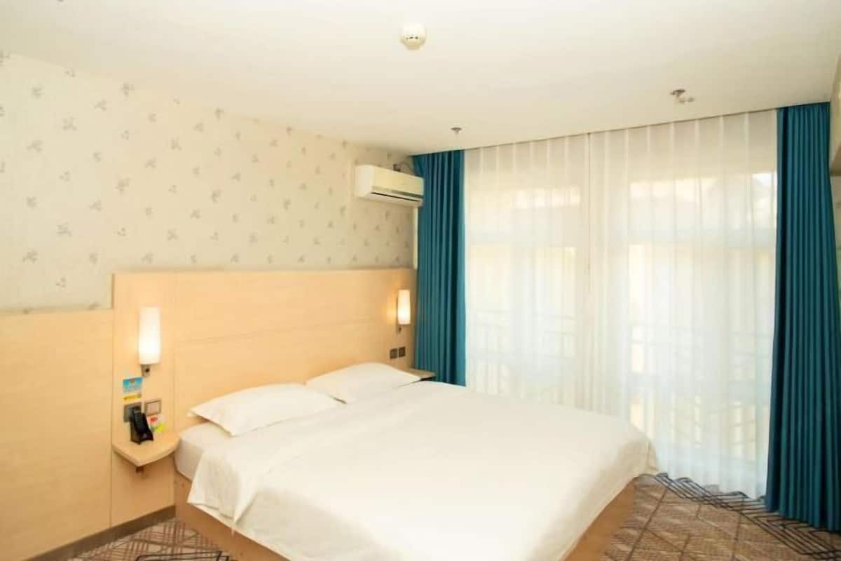 A hotel room with a bed, a nightstand, an air conditioning unit, and a window with curtains.