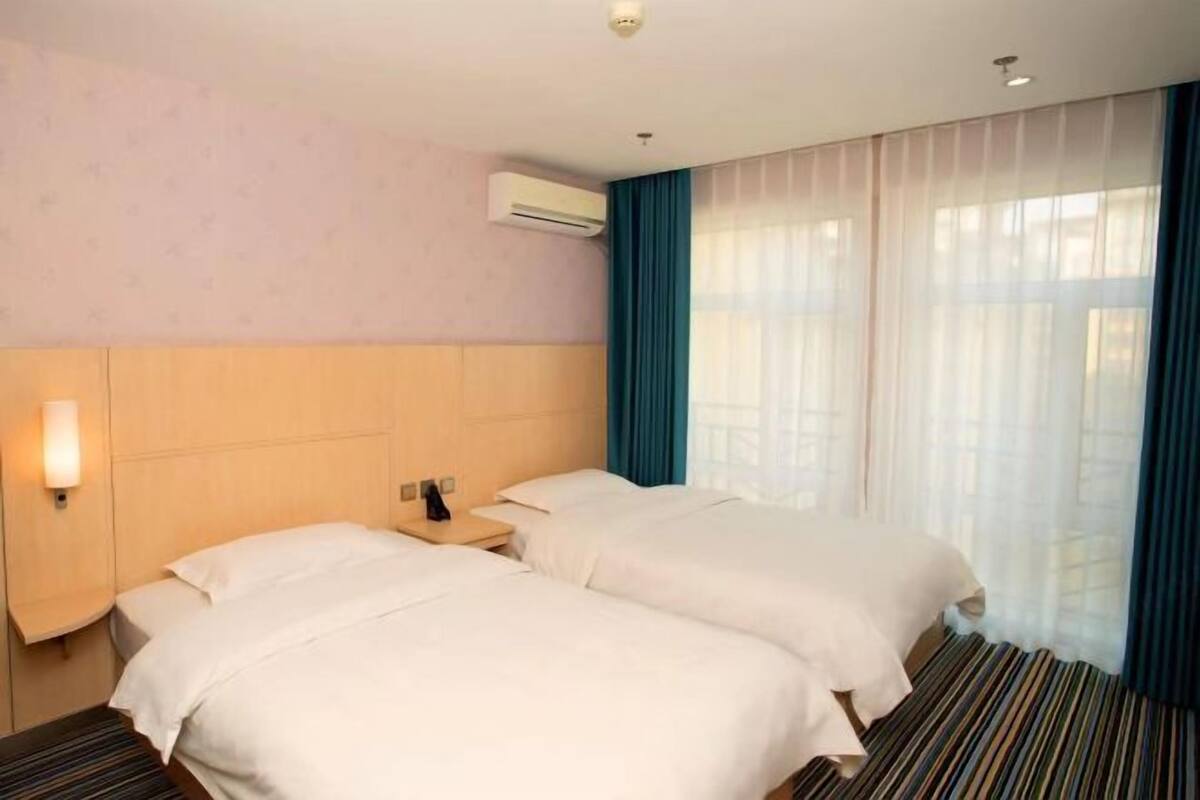 A hotel room with two beds, a nightstand, an air conditioning unit, and a window with sheer curtains.