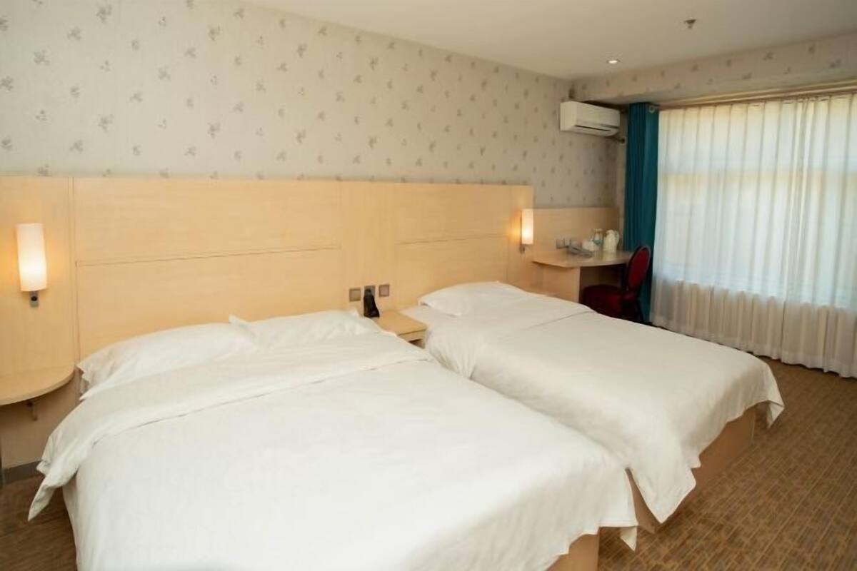 A hotel room with two beds, a desk, a chair, a window with curtains, and an air conditioning unit.