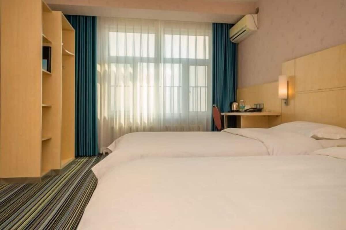 A hotel room with two beds, a desk with a chair, a bookshelf, and a window with curtains.