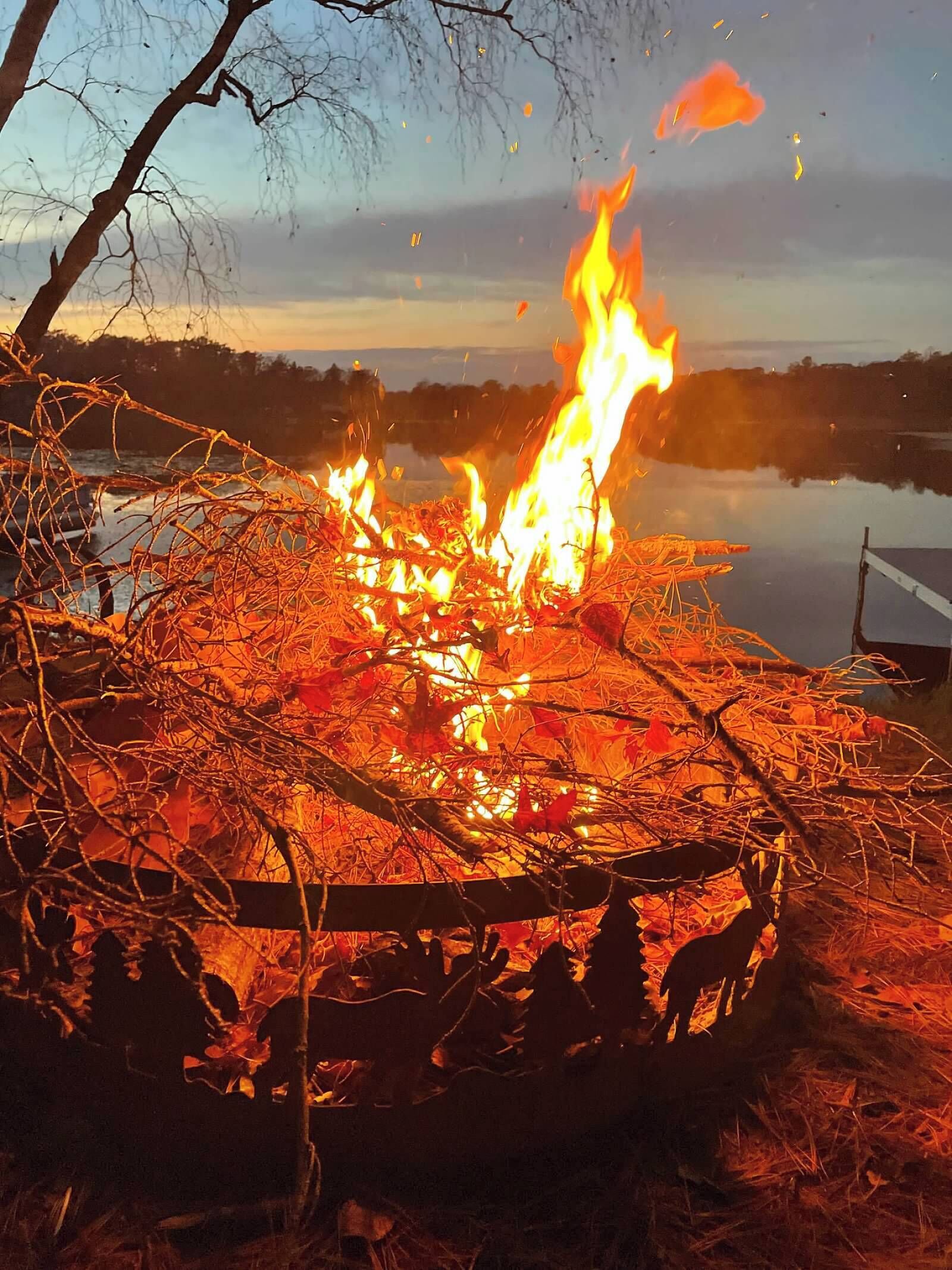 Un feu de camp avec des flammes, entouré de branches et de feuilles sèches, avec une étendue d’eau et des arbres en arrière-plan.