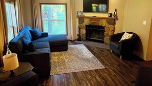 A living room with a stone fireplace, a large flat-screen TV mounted above it, a sectional sofa, a smaller armchair, and a wooden floor. - Spacious cabin right on the lake, perfect for your next family vacation! (Luck)