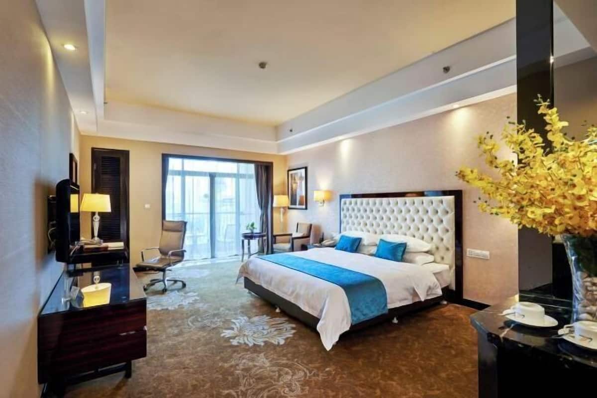A hotel room with a large bed, a desk, a chair, a desk lamp, a vase with flowers, and a view of the outdoors.