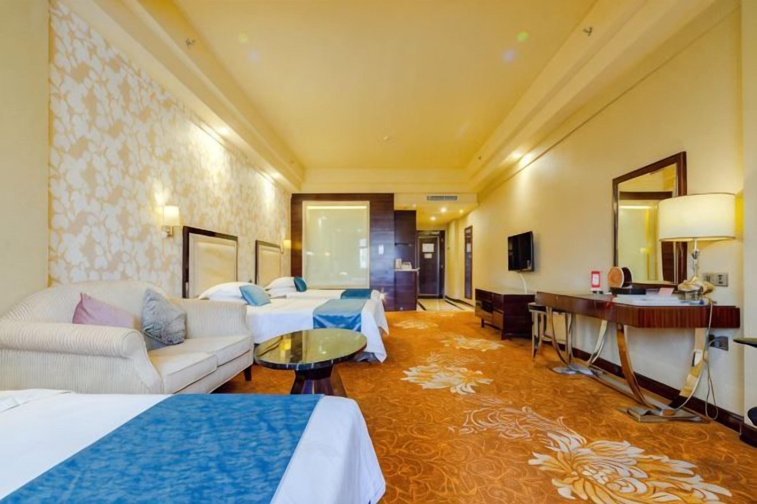 A hotel room with a sofa, two beds, a small table, a mirror, and a carpeted floor.